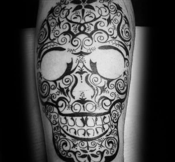 Decorative tribal skull tattoo with intricate swirls, dots, and symmetrical patterns, resembling a sugar skull style on the calf.