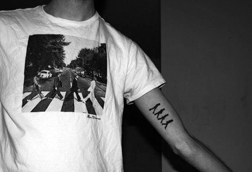 Simple black tattoo on the inner arm showing The Beatles in their Abbey Road walking pose, matching the iconic image on the T-shirt.
