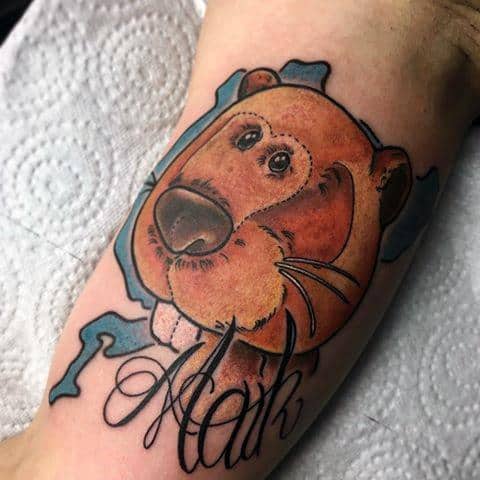 Cartoon-style beaver tattoo peeking through blue background with a sweet expression and the name “Mark” in elegant script below.
