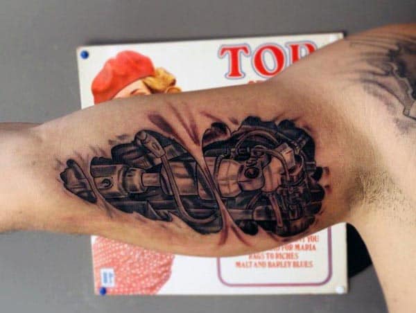 Black and grey biomechanical tattoo on the inner bicep, depicting realistic gears, tubes, and pistons beneath torn skin for a 3D effect