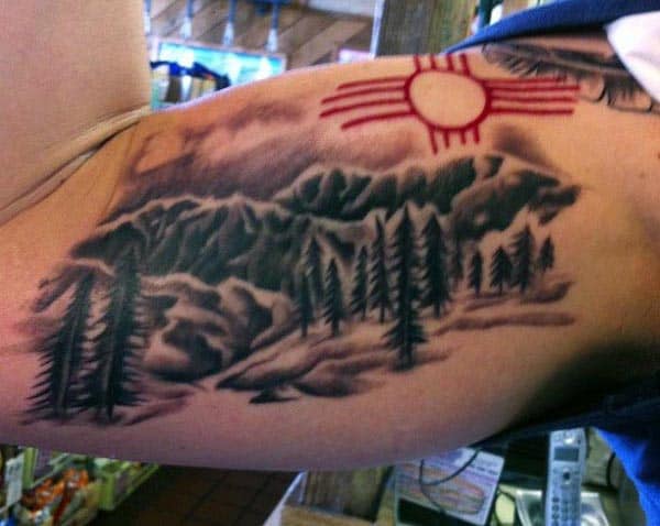 Black and grey mountain landscape tattoo with pine trees, accented by a red Zia sun symbol on the upper inner bicep