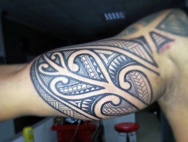 Bold Polynesian tribal tattoo on the upper bicep, featuring intricate black linework, geometric patterns, and swirling shapes