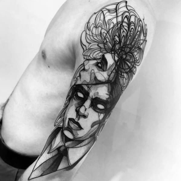 Black and grey surreal tattoo on a man's upper arm featuring a detailed face in a suit, abstract shading, and a large floral design.