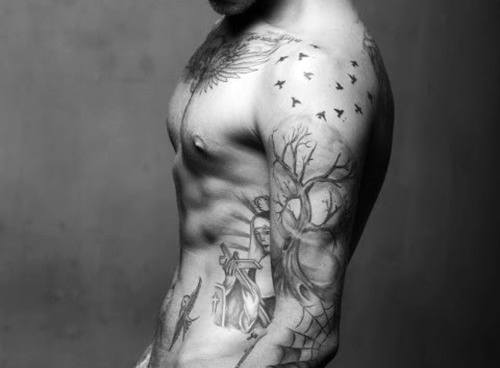 Shirtless man with detailed tattoos of birds, a tree, and a figure on his arm and chest