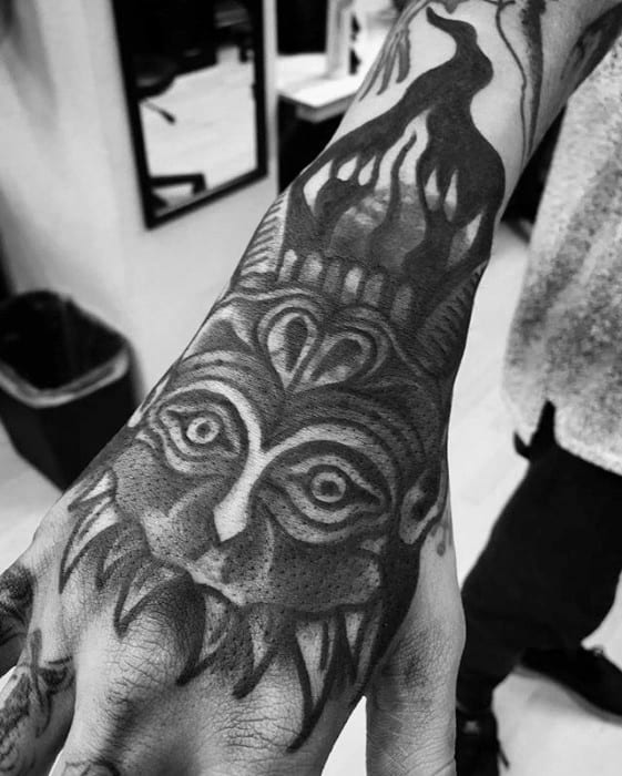 Blast over tattoo on hand of a stylized face with sharp teeth and flame-like designs