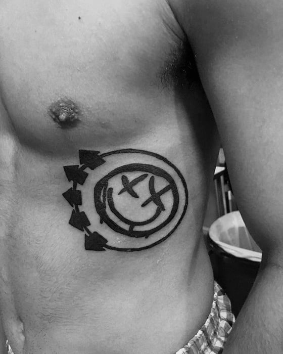 Black and white tattoo of the Blink-182 smiley face logo with arrows, placed on the side of the torso.