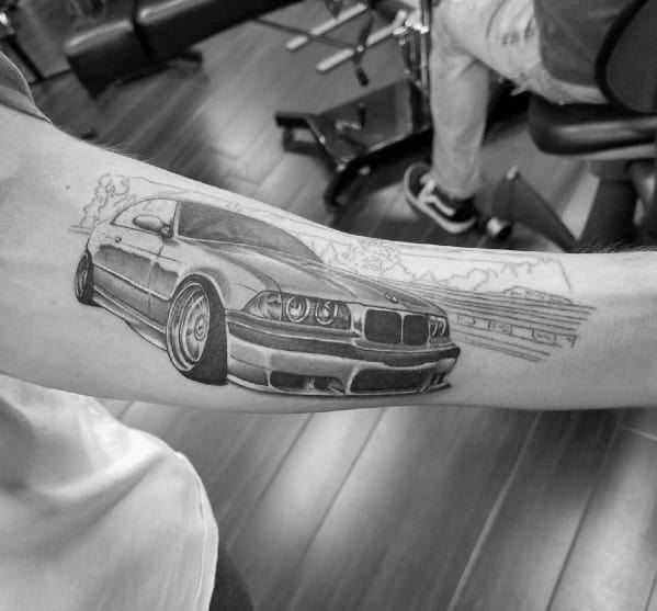 Realistic BMW E39 tattoo on forearm, featuring fine details and a racetrack background for a sleek and dynamic automotive design.