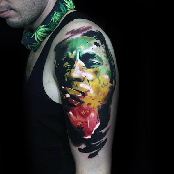 Vibrant upper arm tattoo of Bob Marley with abstract strokes in red, yellow, and green, creating a bold watercolor-style portrait.