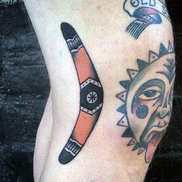 Bold boomerang tattoo with a black and orange color scheme and central geometric symbol, placed on the side of the thigh.