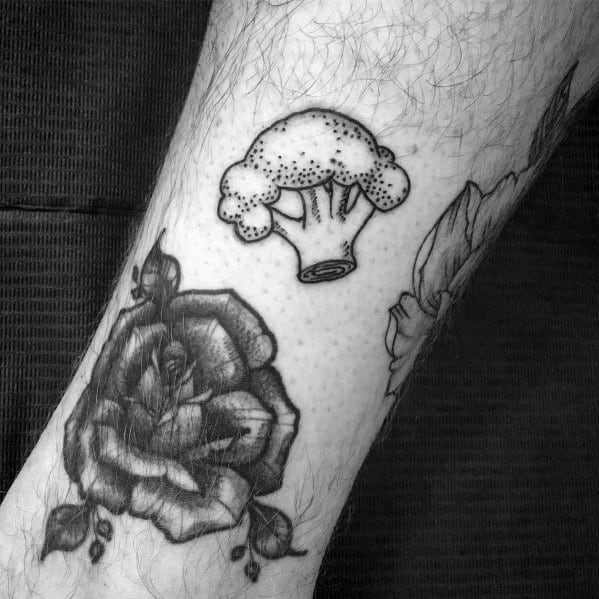 Small black ink broccoli tattoo on the leg, featuring fine dot work and bold outlines, complementing surrounding floral designs for a unique look.