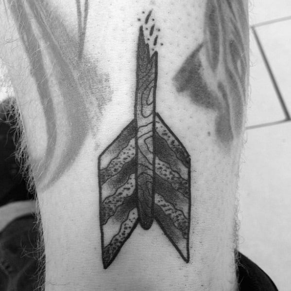Black and grey tattoo of a broken arrow fletching with bold striped pattern, rough edge, and small fragments breaking off the top.