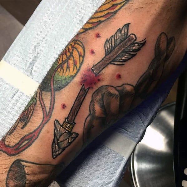 Broken arrow tattoo with realistic detailing and a red burst at the break point, featuring a stone arrowhead and feathered fletching on the forearm.