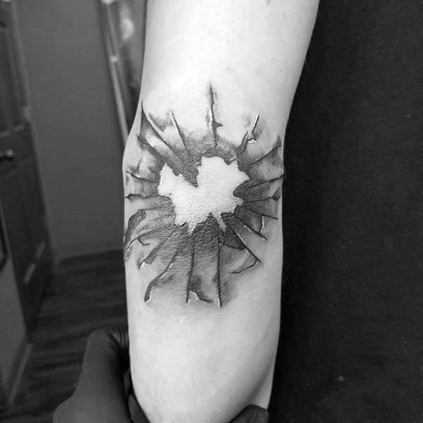 Minimalist black and grey tattoo of a cracked bullet hole, centered on the forearm with radiating fracture lines for a realistic shattered effect.