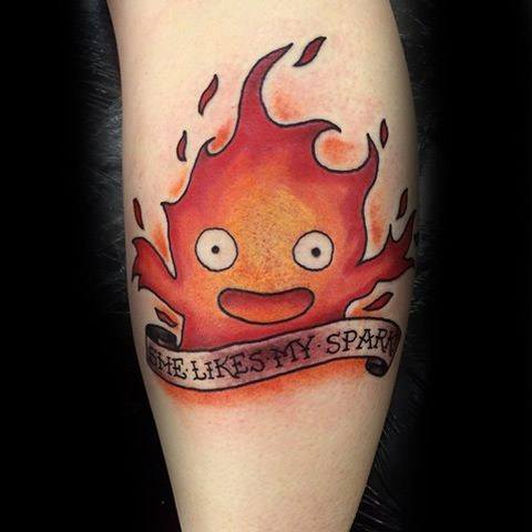 Cheerful Calcifer tattoo with bright flames, a smiling face, and a scroll below reading “She Likes My Spark.”