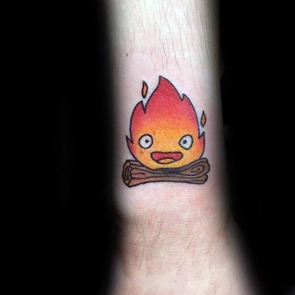 Small Calcifer tattoo with a happy expression, sitting on neatly drawn logs, and glowing red-orange flames on the forearm.