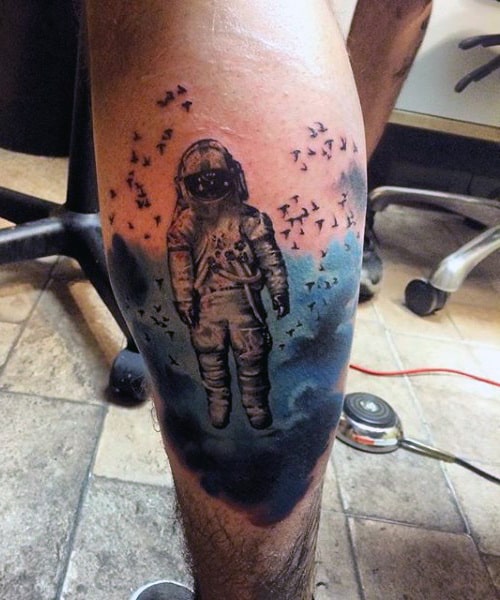 Calf tattoo of an astronaut floating in a cosmic scene, surrounded by a dark blue nebula and a flock of black birds for a surreal effect