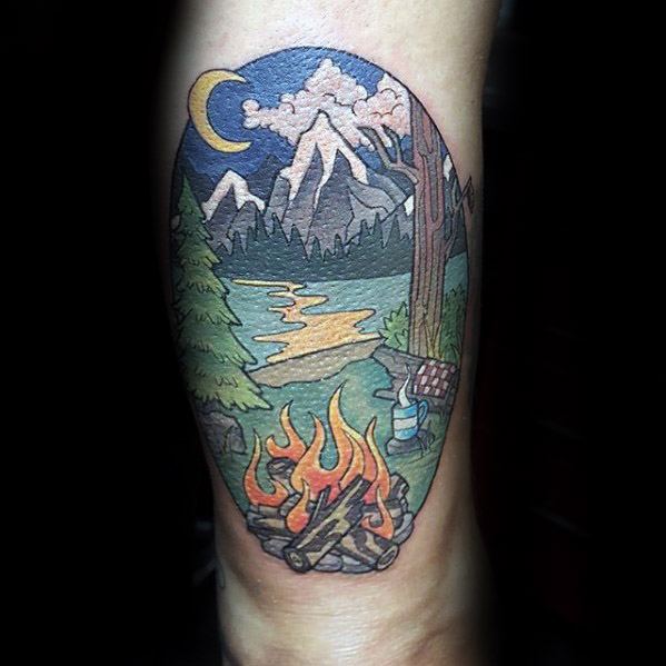 Colorful tattoo of a cozy campsite with a campfire, river, mountains, crescent moon, and trees in a cartoon-style oval frame.