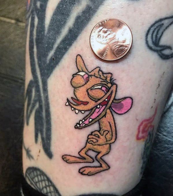 Small and detailed tattoo of Ren from Ren & Stimpy with a crazed expression, positioned next to a penny for scale on a heavily tattooed leg.