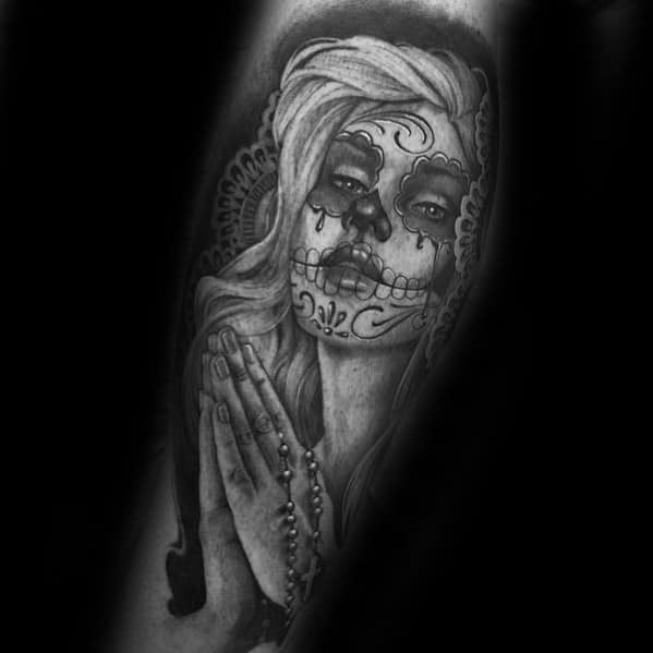 La Catrina tattoo of a woman with sugar skull makeup in prayer, holding a rosary, with detailed hair and lace-like background.