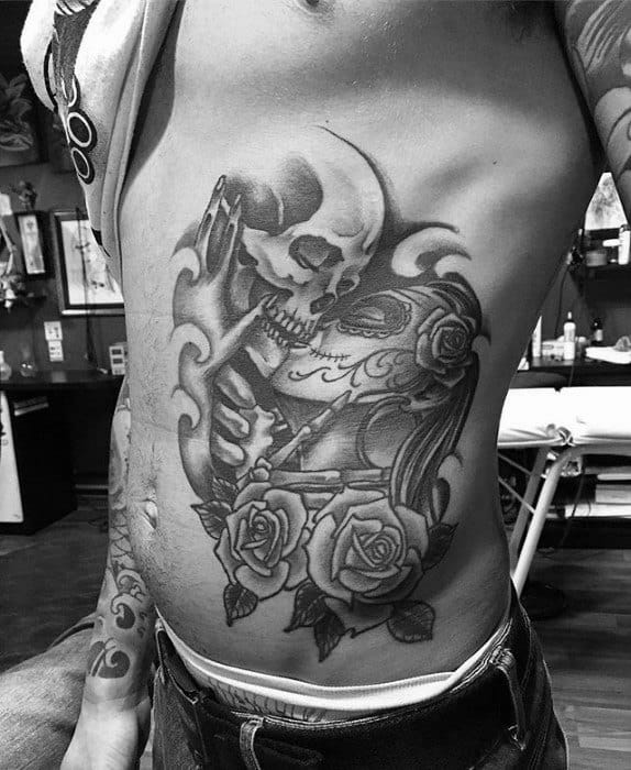 La Catrina rib tattoo of a skull kissing a woman with sugar skull makeup, surrounded by roses, smoke, and bold black shading.