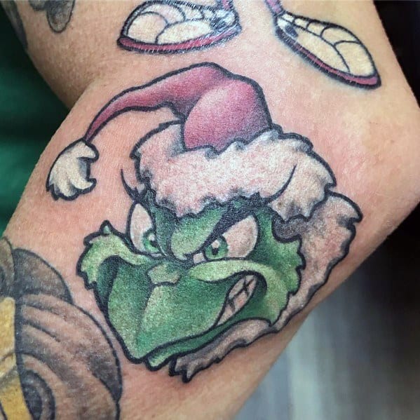 Christmas tattoo of the Grinch wearing a Santa hat, smirking mischievously, with bold outlines and shading for a classic holiday cartoon look.