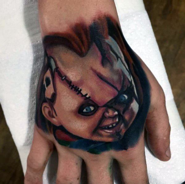 Realistic Chucky hand tattoo with vivid colors, featuring his sinister grin, stitched face, and piercing blue eyes for a bold horror-inspired design.