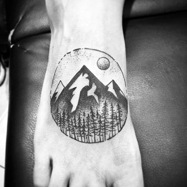 Black and white tattoo on foot showing a mountain scene with trees and a sun or moon in a circular design