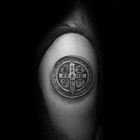 Realistic tattoo of a Saint Benedict medal on the upper arm, featuring intricate details and shading for a three-dimensional effect.