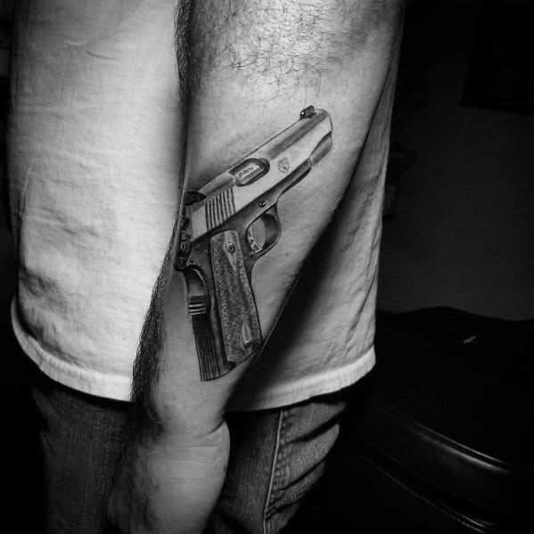Realistic black and gray tattoo of a 1911 pistol on the outer forearm, featuring fine details and shading for a bold and three-dimensional effect.