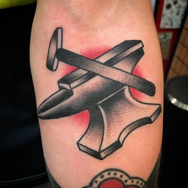 Bold anvil and hammer tattoo on the forearm with classic black shading and a red background glow, creating a strong and timeless look.