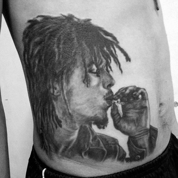 Black and gray rib tattoo of Bob Marley in profile, holding and smoking a joint, with textured dreadlocks and a calm, focused expression.