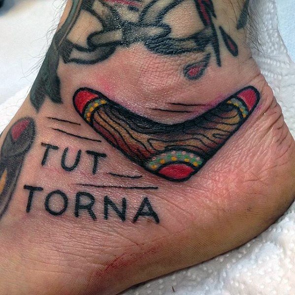 Boomerang tattoo on the side of the foot with earthy colors, motion lines, and the phrase TUTTO TORNA in bold black letters.
