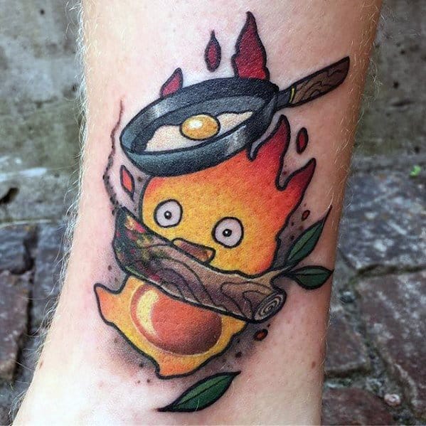 Lively Calcifer tattoo with a frying pan and egg above, expressive eyes, a log in front, and green leaves accenting the flames.