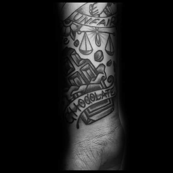 This black and grey tattoo features a **chocolate bar with a "CHOCOLATE" banner**, integrated into a larger design with scales and symbols. 🍫