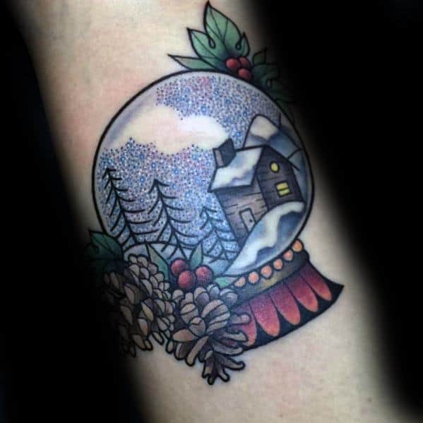 Christmas tattoo of a festive snow globe featuring a cozy cabin, snow-covered trees, pinecones, holly, and red berries for a classic holiday design.