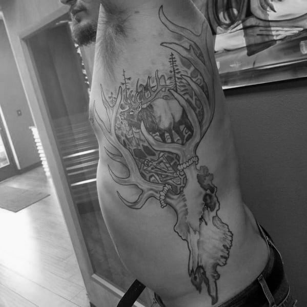Black and grey elk skull tattoo on the ribcage, featuring intricate antlers with a detailed forest and elk scene integrated into the design.