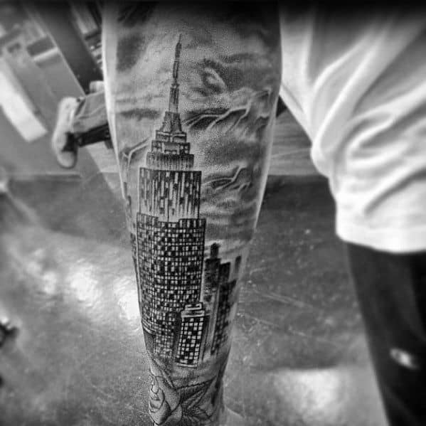 Detailed Empire State Building tattoo showcasing the NYC skyline against a dramatic, cloudy background, inked on a man's forearm.