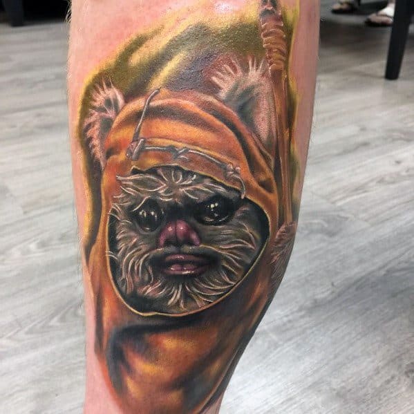 Realistic Ewok tattoo with rich shading and lifelike fur, wearing a detailed orange hood and holding a spear against a soft forest backdrop.