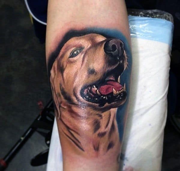 Realistic Golden Retriever tattoo on the forearm, featuring vibrant colors, detailed fur shading, and a joyful expression against a blue background.