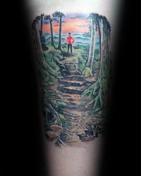 This tattoo illustrates a hiker in a red shirt standing on a scenic forest trail at sunset, surrounded by lush greenery and detailed roots.