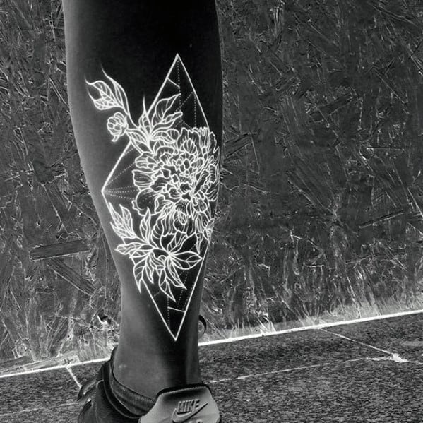 Inverted floral tattoo of detailed peony blooms within a diamond frame, glowing white against a dark leg background.