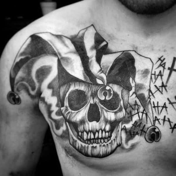 A black and gray tattoo of a skull wearing a jester hat, with dramatic shading and smoke effects, placed on the chest. The background features abstract marks and symbols.