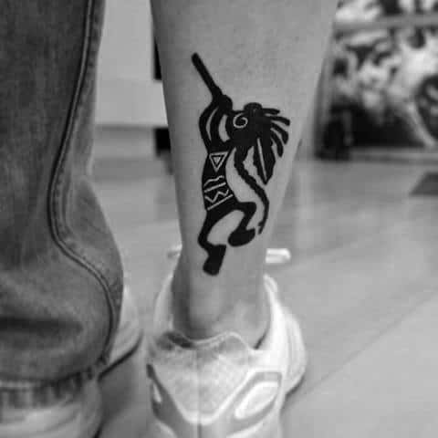 Bold black Kokopelli tattoo with tribal geometric patterns on the body and flute, placed on the lower leg.