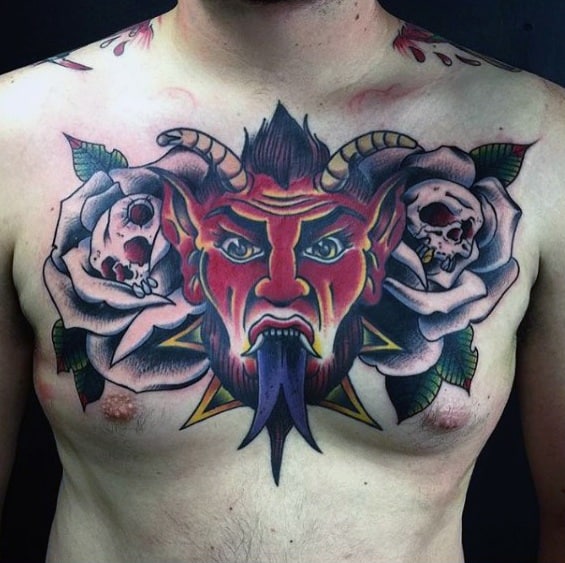 Bold Krampus chest tattoo with a red demon face, yellow horns, long blue tongue, flanked by skull-filled roses and dark green leaves.