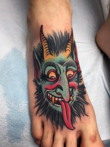 Colorful Krampus tattoo on foot featuring a blue-green face, yellow horns, red eyes and tongue, exaggerated features, and wild black hair.