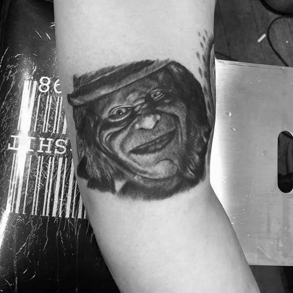 Black and grey leprechaun tattoo with a sinister smile, shadowed eyes, and a tilted hat, giving a creepy and haunting expression.