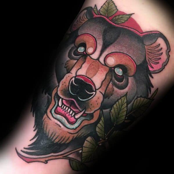 Neo-traditional bear tattoo featuring a stylized bear with glowing eyes, a red hat, and detailed leaves surrounding its face.