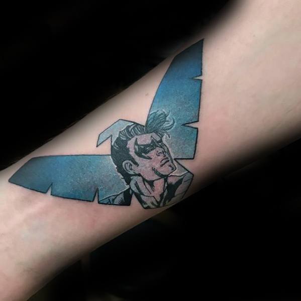 Nightwing forearm tattoo blending comic-style portrait within a blue winged emblem, giving a sharp and sleek visual effect.