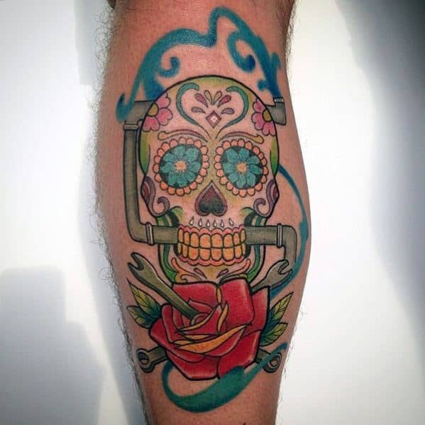 Colorful tattoo of a sugar skull made from plumbing pipes, decorated with flowers and swirls, with a red rose and wrench below.
