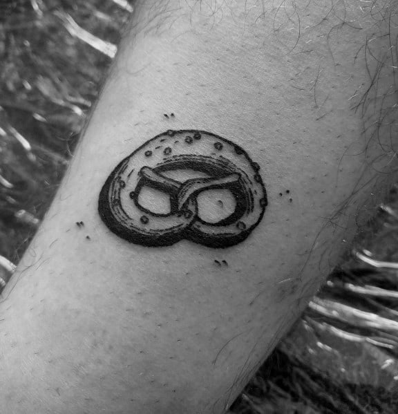Small black and grey pretzel tattoo with detailed shading and salt specks, creating a realistic and textured look on the skin.